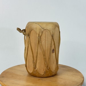 May include: A cylindrical wooden drum featuring a light tan natural wood grain body. The top and bottom edges are covered with stretched tan rawhide, which is laced together in a crisscross pattern around the sides using thin rawhide strips.