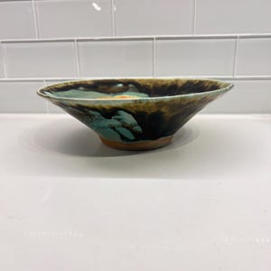May include: A handmade ceramic bowl with a wide, flared shape. The interior features a blend of turquoise, brown, and black glaze, creating a marbled effect. The exterior is a natural, unglazed clay color.