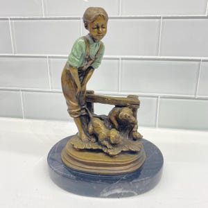 May include: A bronze-toned statue of a young person in overalls and a light green shirt, looking down at two puppies. The statue is on a round, black marble base. The puppies are near a wooden fence.