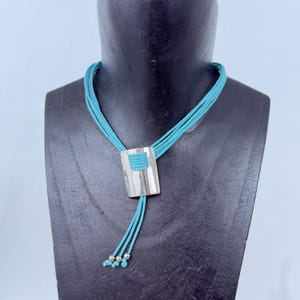 May include: A turquoise necklace with a silver rectangular pendant. The pendant has a turquoise cord wrapped around the center. The necklace has multiple turquoise cords and small silver beads at the ends.