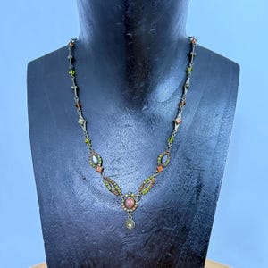 May include: A silver necklace with a variety of colourful beads and gemstones. The necklace features oval and diamond-shaped accents with orange, green, and clear stones. A central pendant with a round, dangling charm completes the design.