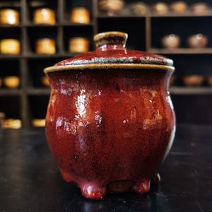 May include: A small, round, lidded ceramic jar with a glossy, deep red glaze featuring dark brown speckles and a textured finish. The jar stands on three short, rounded feet and includes a matching lid with a small, knob-like handle on top.