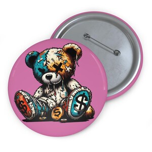 Könnte beinhalten: Ein pinkfarbener Button mit einem Cartoon-Teddybären mit einem Patchwork-Design in Blau, Orange und Weiß. Der Bär hat Dollarzeichen auf seinen Füßen und Münzen in der Nähe. Ein zweiter Button mit Anstecknadel ist sichtbar.