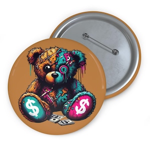 Könnte beinhalten: Ein runder Knopf mit einem Cartoon-Teddybär-Design. Der Bär ist mehrfarbig mit Dollarzeichen auf seinen Pfoten und einem tropfenden Farbeffekt. Der Knopf befindet sich auf einem tanfarbenen Hintergrund mit einem silbernen Nadelrücken.
