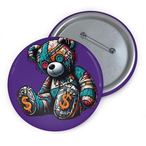 Könnte beinhalten: Ein violetter Button mit einem Cartoon-Teddybär-Design. Der Bär ist türkis, orange und braun mit Dollarzeichen auf den Füßen. Der Button hat eine silberne Rückseite.