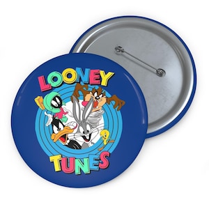 Peut inclure: Un bouton rond bleu avec des personnages de Looney Tunes. Le bouton a une épingle argentée au dos. Le logo Looney Tunes est en haut et en bas en texte coloré. Les personnages incluent Bugs Bunny, Daffy Duck et Taz.