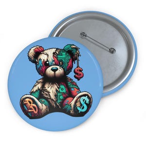 Könnte beinhalten: Ein runder, hellblauer Button mit einer bunten Illustration eines Teddybären mit Dollarzeichen. Der Bär ist mit verschiedenen Mustern und Farben geflickt, darunter Rot, Grün und Blau. Ein zweiter Button ist im Hintergrund sichtbar.