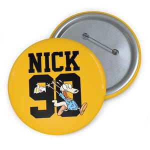 Peut inclure: Un bouton jaune avec le mot "NICK" en lettres noires au-dessus du chiffre "99". Le bouton présente une image de dessin animé d'un personnage ressemblant à un chien avec une expression surprise. Le bouton a un dos à épingle en métal.
