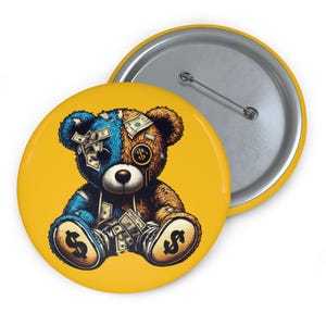 Könnte beinhalten: Ein Button-Pin mit einem Cartoon-Teddybär-Design. Der Bär ist in blaue und braune Hälften geteilt und mit Geldscheinen verziert. Der Bär sitzt auf einem gelben Hintergrund mit silberner Nadelrückseite.