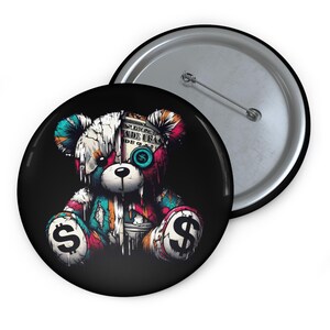 Könnte beinhalten: Ein Ansteckbutton mit einem farbenfrohen, künstlerischen Design eines Teddybären. Der Bär hat ein Dollarzeichen auf jeder Pfote und ein Dollarzeichen für ein Auge. Der Text "MADE USA" ist auf dem Kopf des Bären zu sehen. Der Button ist schwarz mit silberner Rückseite.