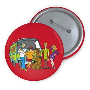 May include: A red button featuring the Scooby-Doo gang and their van, the Mystery Machine. The cartoon characters are in front of the van. The button has a silver pin on the back.