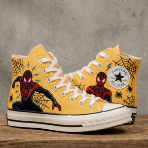 May include: Yellow high-top Converse sneakers featuring Spider-Man graphics. The sneakers have white laces, a white rubber toe cap, and a black sole. Spider-Man is depicted in action with spiderwebs and the Converse All Star logo.