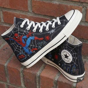 May include: Black high-top sneakers featuring Spider-Man embroidery and a web design. The sneakers have white laces, a white sole, and a Converse All Star logo patch. The shoes are made of canvas.