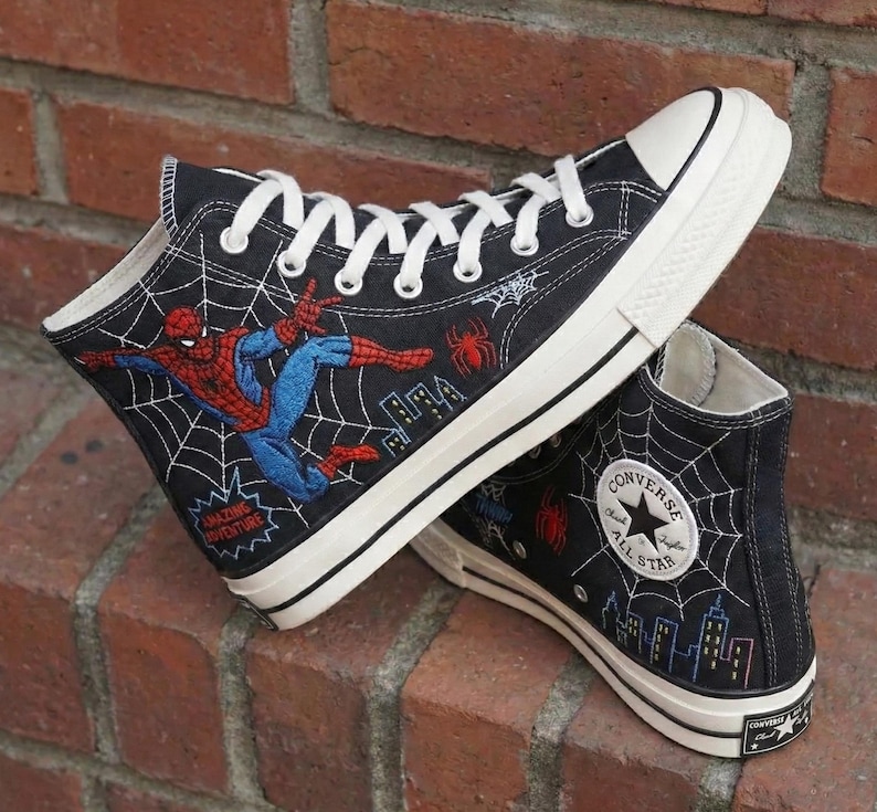 May include: Black high-top sneakers featuring Spider-Man embroidery and a web design. The sneakers have white laces, a white sole, and a Converse All Star logo patch. The shoes are made of canvas.