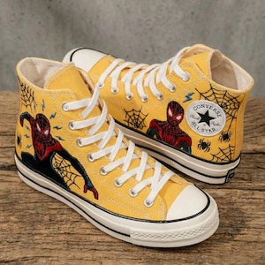 May include: A pair of yellow high-top canvas sneakers featuring a Spider-Man design. The sneakers have white laces, a white toe cap, and a black sole. The Spider-Man graphic includes webs, lightning bolts, and the Converse All Star logo.