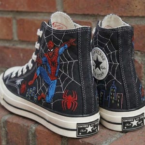 May include: Black high-top Converse sneakers featuring Spider-Man and a spiderweb design. The sneakers have a white rubber sole and laces, with the Converse All Star logo on the side and heel. The Spider-Man graphic is in red, blue, and white.