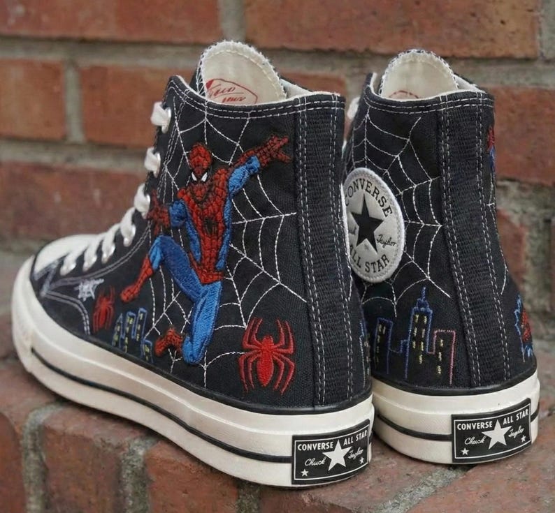 May include: Black high-top Converse sneakers featuring Spider-Man and a spiderweb design. The sneakers have a white rubber sole and laces, with the Converse All Star logo on the side and heel. The Spider-Man graphic is in red, blue, and white.