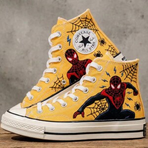 May include: Yellow high-top Converse All Star sneakers featuring Spider-Man graphics. The sneakers have white laces, a black and white sole, and a Spider-Man logo patch. Spiderwebs and lightning bolts are also featured on the canvas.