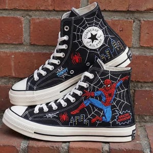 May include: Black high-top Converse sneakers featuring Spider-Man graphics. The sneakers have white laces, soles, and toe caps. Designs include spiderwebs, spiders, cityscapes, and the text "THWIP!" and "AMAZING ADVENTURE".