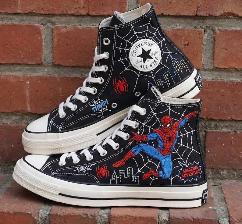 May include: Black high-top Converse sneakers featuring Spider-Man graphics. The sneakers have white laces, soles, and toe caps. Designs include spiderwebs, spiders, cityscapes, and the text "THWIP!" and "AMAZING ADVENTURE".