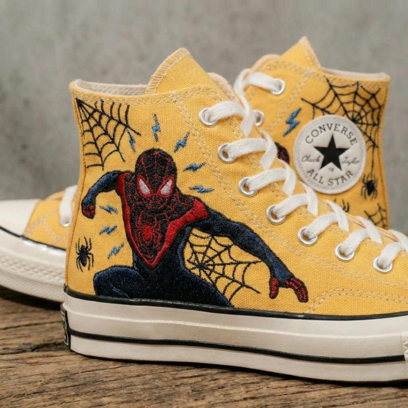 May include: Yellow high-top sneakers featuring a Miles Morales Spider-Man design. The sneakers have white laces, a black and white sole, and a Converse All Star logo. Embroidered spiderwebs and lightning bolts accent the design.