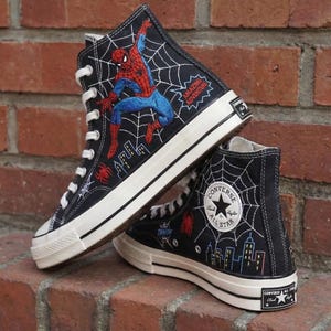 May include: Black high-top sneakers featuring a Spider-Man design. The sneakers have a white sole and laces, with embroidered images of Spider-Man, a spider web, and city skyline. The text "AMAZING ADVENTURE" is also visible.