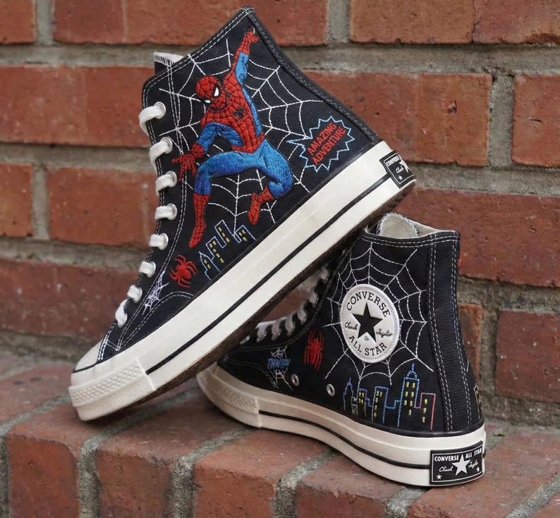 May include: Black high-top sneakers featuring a Spider-Man design. The sneakers have a white sole and laces, with embroidered images of Spider-Man, a spider web, and city skyline. The text "AMAZING ADVENTURE" is also visible.