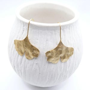Peut inclure: Une paire de boucles d'oreilles pendantes dorées en forme de feuilles de ginkgo. Chaque boucle présente une surface en métal martelé texturé et un crochet fin et incurvé. Le design reproduit la silhouette en éventail d'une feuille.