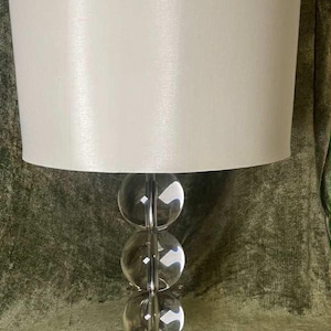 Puede incluir: Lámpara de mesa decorativa con una pantalla blanca y una base compuesta por tres esferas de vidrio transparente apiladas verticalmente. La base está soportada por un soporte de metal plateado. La lámpara está diseñada para uso en interiores.