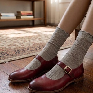 Può includere: Un paio di scarpe Mary Jane in pelle bordeaux con cinturino e fibbia dorata. Le scarpe sono abbinate a calzini grigi a coste. Le scarpe sono su un pavimento in legno.