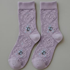 May include: A pair of lavender socks with an intricate floral lace pattern. The socks feature a detailed design with small blue and gray flower accents, adding a touch of elegance to the overall look. The socks are made of a soft, comfortable material.