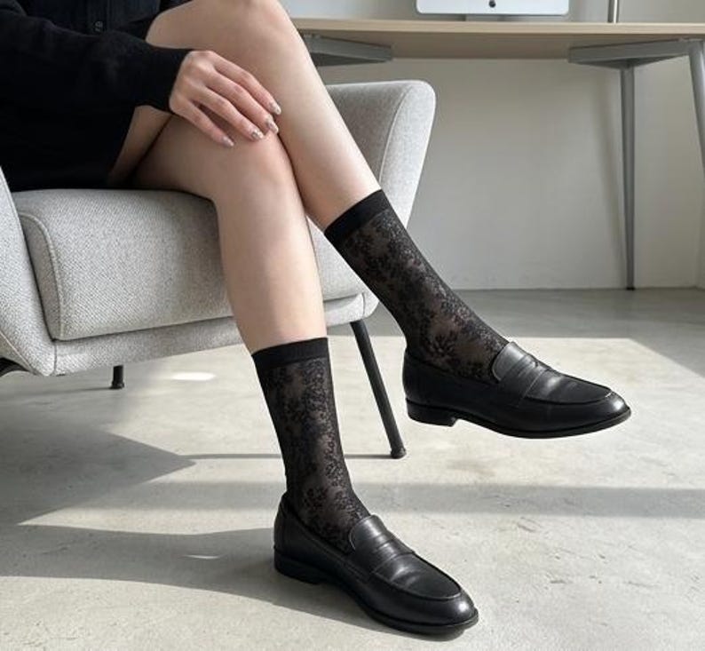 May include: Black leather loafers with a classic design. The shoes are paired with sheer black socks featuring a floral pattern. The socks are visible above the shoes, creating a contrast in texture and style. The overall look is simple and elegant.