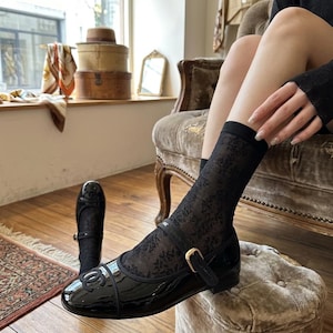 May include: Black patent leather Mary Jane flats with a gold buckle and a decorative logo. The shoes are paired with black lace socks. The image is taken indoors, with a vintage aesthetic.
