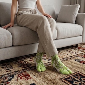 May include: A person wearing white and green patterned socks with a split toe design. The socks are worn with beige trousers while sitting on a grey sofa. The socks have a light green toe and a white background with a green floral pattern.