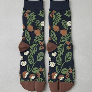 May include: Navy blue tabi socks with a botanical design. The pattern includes green leaves, brown acorns, and white seed shapes. The toe and cuff are brown. A unique sock style.