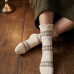 May include: Cream-coloured socks with a patterned design in green and purple. The socks have a lace trim at the top and are displayed on a wooden floor. These socks are a cosy and comfortable accessory.
