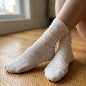 May include: A pair of off-white, patterned socks. The socks are made of a soft material and have a delicate, openwork design. The socks are slightly scrunched down around the ankles. A gold anklet is visible.