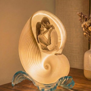 May include: A seashell-shaped lamp with a photo of a couple embracing inside. The lamp is white with a blue wave-like base. The lamp is lit, creating a warm glow.