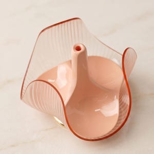 May include: A pink and clear decorative vase with a unique, curved design. The vase features a central, upright spout and a wide, fluted rim. The base has a gold accent. The vase is made of glass and is suitable for home decor.