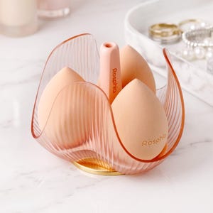 May include: A clear, pink, petal-shaped makeup sponge holder with a gold base. The holder contains three peach-colored makeup sponges and a pink sponge applicator. The text "Rosphia" is visible on the applicator and one sponge.