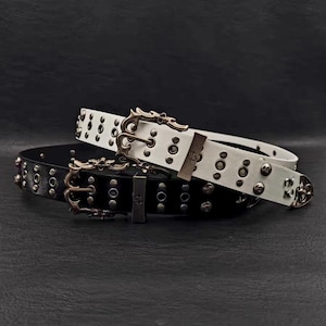 Puede incluir: Dos cinturones de cuero, uno negro y otro blanco, con hebillas de metal ornamentadas y detalles tachonados. Los cinturones están apilados, mostrando sus colores contrastantes y su hardware único.