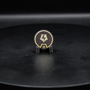 May include: A circular dark brown token with a serrated edge, featuring a light-colored lightning bolt icon centered within a pentagonal shape and the number 11 printed below it, held upright by a small black stand.