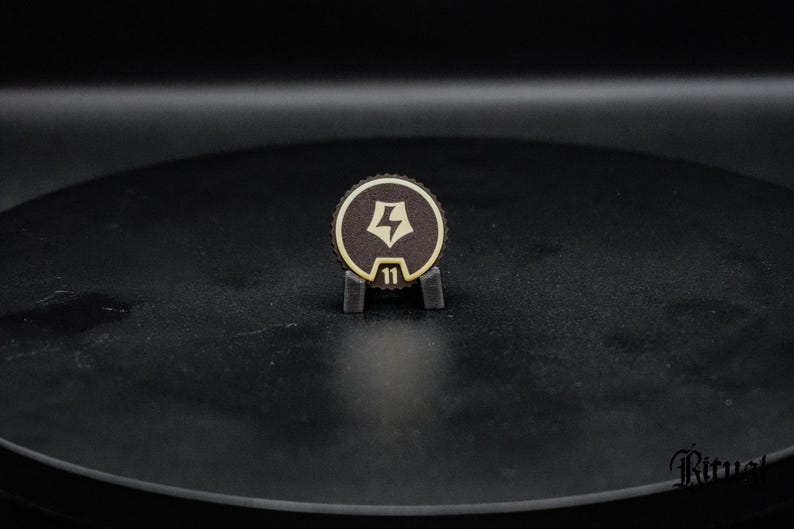 May include: A circular dark brown token with a serrated edge, featuring a light-colored lightning bolt icon centered within a pentagonal shape and the number 11 printed below it, held upright by a small black stand.