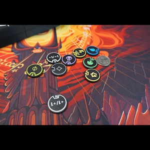 May include: A collection of small, round, black game tokens with colored edges and various white or colored icons and numbers printed on them, including symbols for storm, skull, tree, and +1/+1 counters, alongside a silver US quarter coin.