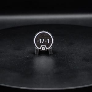 May include: A circular black token with a white border and white text reading -1/-1 and the number 15 at the bottom. The token is held upright by a small black stand. The design features a textured surface and a notched edge around the perimeter.
