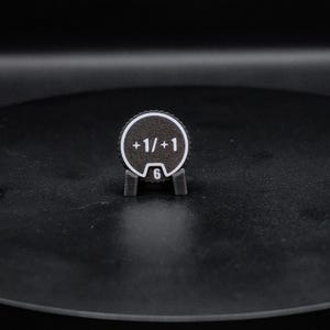 May include: A circular black token with a white border and white text reading +1/+1. The token features a small notch at the bottom containing the number 6 in white. It is held upright by a small, dark gray, two-pronged plastic stand.