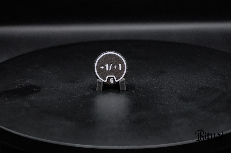 May include: A circular black token with a white border and white text reading +1/+1. The token features a small notch at the bottom containing the number 6 in white. It is held upright by a small, dark gray, two-pronged plastic stand.