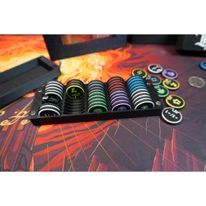 Könnte beinhalten: Ein schwarzes Kunststoff-Aufbewahrungstablett mit fünf Stapeln runder Spielmarken. Die Marken zeigen verschiedene farbige Symbole, darunter einen gelben Blitz, einen grünen Baum, ein rotes Symbol, einen violetten Totenkopf und eine blaue Sonne.