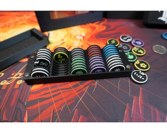May include: A black plastic storage tray containing five stacks of circular game tokens. The tokens feature various colored icons including a yellow lightning bolt, green tree, red symbol, purple skull, and blue sun. Additional loose tokens are nearby.