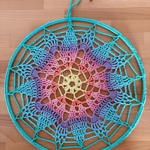 May include: A handmade dreamcatcher with a turquoise outer ring and a colorful web design. The web features a gradient of colors, including yellow, pink, purple, and blue. The dreamcatcher is designed to be hung and is displayed on a wooden surface.